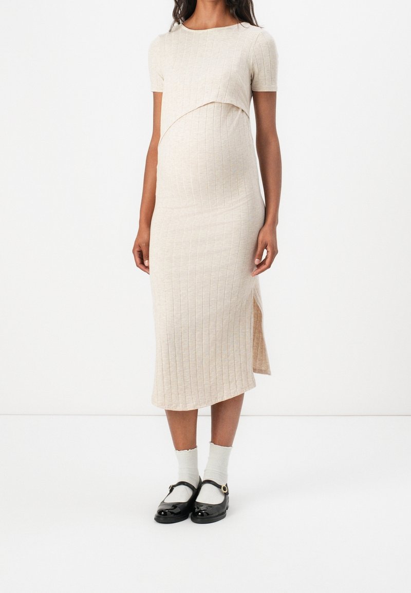 Robe de maternité en maille côtelée beige clair avec manches courtes, sur-couche courbée au niveau de la poitrine et fente latérale, portée avec des chaussures noires et des chaussettes blanches.