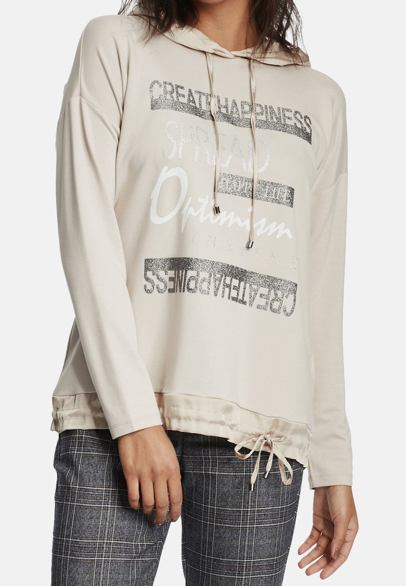 Kvinna klädd i en beige huvtröja med dragsko och glittrande text om lycka och optimisme, i kombination med grå rutiga byxor.