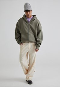 Un sweat à capuche zippé vert olive avec des poches avant, associé à un pantalon large crème et un bonnet en tricot gris clair. Des chaussures noires complètent le look.