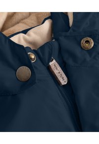 Marineblaue wasserdichte Jacke mit Fleece-Futter, ausgestattet mit bronzefarbenen Knöpfen und einem Reißverschluss-Zipper mit eingeprägtem Branding. Weicher Texturdetail sichtbar.