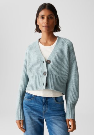 Donna che indossa un cardigan a maglia grosso di colore azzurro chiaro con bottoni, sopra una camicia bianca e jeans blu, in piedi contro uno sfondo neutro.
