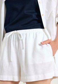 Shorts bianchi in lino con elastico in vita e coulisse. Presentano tasche laterali e un tessuto leggero e texture. Abbinati a una maglietta blu navy.