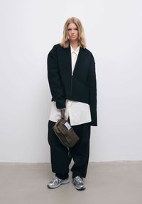 Sort ribbet jakke med lynlås foran, båret over en hvid skjorte, parret med løse sorte bukser og sølvfarvede sneakers. Olivengrøn taske med lynlås.