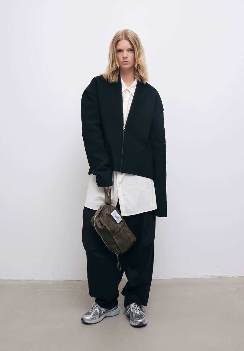 Sort ribbet jakke med lynlås foran, båret over en hvid skjorte, parret med løse sorte bukser og sølvfarvede sneakers. Olivengrøn taske med lynlås.