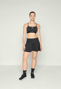 Μαύρο αθλητικό στηθόδεσμο με λεπτές τιράντες, συνδυασμένο με μια μαύρη φούστα σε φαρδύ στυλ. Και τα δύο κατασκευασμένα από ελαφρύ ύφασμα, με το λογότυπο της Adidas στον στηθόδεσμο.