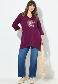 Lila Tunika mit abgerundetem Saum, versehen mit einem goldenen und pinken Grafikdesign, kombiniert mit blauen weiten Jeans und einer gewebten beigefarbenen Tragetasche.