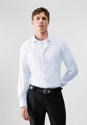 Jeune homme debout, main derrière le dos, portant une chemise blanche boutonnée sur un maillot blanc et un pantalon noir avec une ceinture noire.