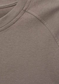 Tessuto color marrone chiaro, texturizzato, con scollo a giro e design delle maniche raglan. Materiale morbido con dettagli di cucitura visibili.