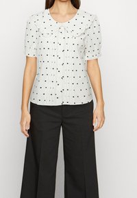 Vit blus med svarta prickar, med en stor rund krage och korta puffärmar, kombinerad med vida svarta byxor.