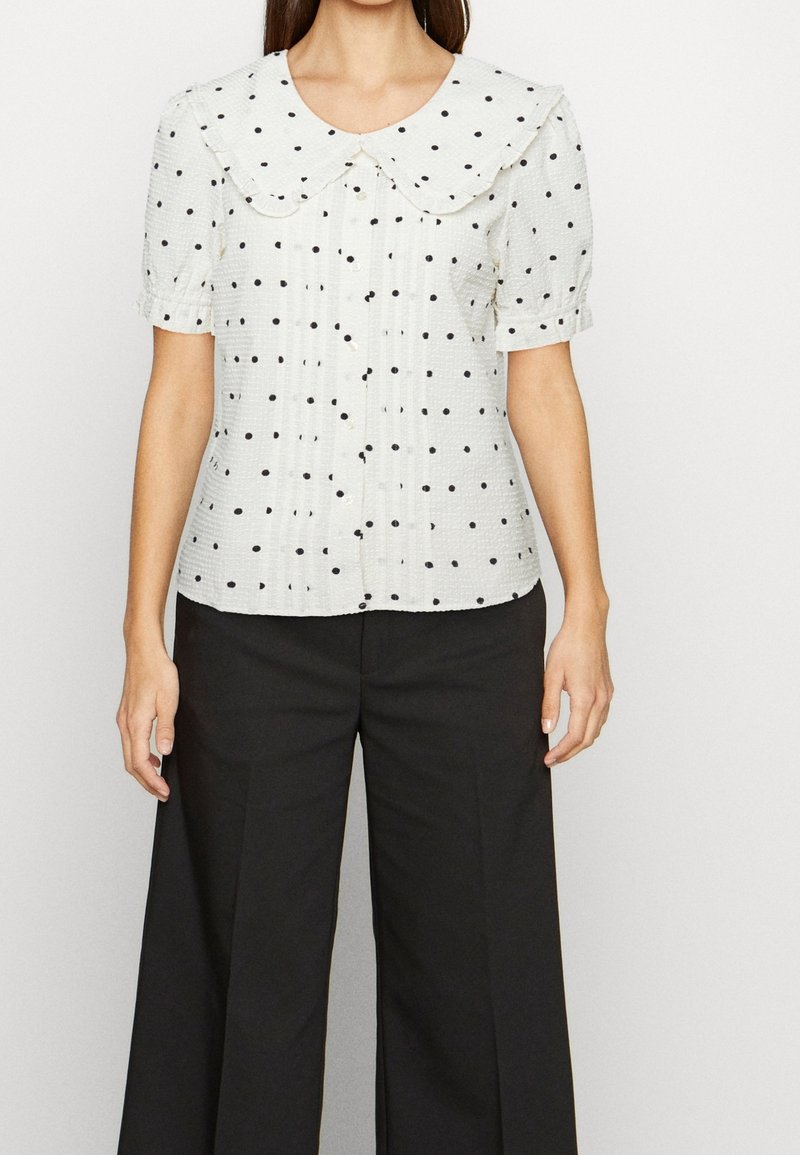 Vit blus med svarta prickar, med en stor rund krage och korta puffärmar, kombinerad med vida svarta byxor.