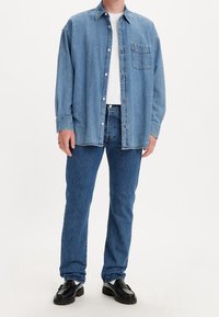 Person wearing blue denim shirt and jeans, white undershirt, and black leather loafers, standing against a white background.