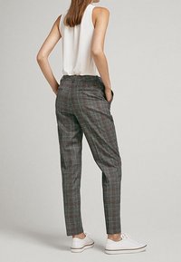 Femme portant un pantalon à carreaux gris, un débardeur blanc et des baskets blanches, se tenant avec les mains dans les poches, face légèrement détourée.