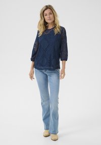 Blusa de encaje azul marino con mangas tres cuartos, que presenta un patrón circular. Combinada con unos vaqueros acampanados de color azul claro y zuecos beige.