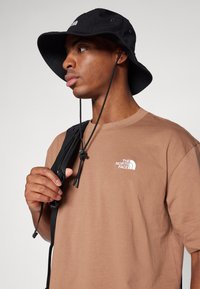 Un chapeau seau noir avec des oeillets, porté par-dessus un t-shirt marron avec un logo blanc. La personne tient une sangle de sac à dos noir.