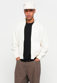 Giacca bomber bianca con colletto alto, cerniera e due tasche frontali. Indossata sopra una maglietta nera e pantaloni a righe marroni. Cappello leggero in testa.