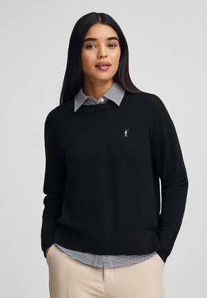 Maglione nero a maglia con una vestibilità rilassata, scollo rotondo e un piccolo logo rosa. Indossato sopra una camicia a colletto a quadri blu e bianchi.