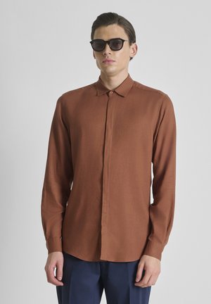 Joven con gafas de sol negras, camisa marrón de manga larga abotonada y pantalones azul oscuro, de pie frente a un fondo claro y liso.