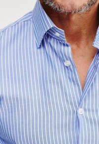 Jasnoniebieska koszula zapinana na guziki w pionowe białe paski. Posiada klasyczny kołnierzyk, małe guziki oraz gładką, bawełnianą fakturę.