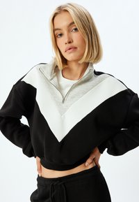 Sudadera recortada en blanco y negro con un diseño en forma de V, que presenta un alto cuello gris y cierre de cremallera. Textura de tela suave y lisa.