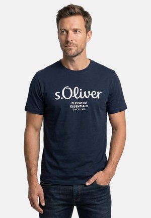 Mężczyzna o krótkich brązowych włosach, ubrany w granatowy t-shirt s.Oliver z białym napisem, stoi z jedną ręką w kieszeni na jednolitym tle.