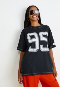 Smiling woman with long braids wears oversized black T-shirt with "95," large sunglasses, hoop earrings, and bright orange pants against white wall.