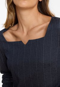 Frau trägt ein navyblaues Oberteil mit Nadelstreifen und einem einzigartigen quadratischen Ausschnitt, der eine kleine mittige Einkerbung aufweist und die Schlüsselbeine zeigt.