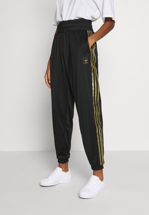 Personne portant un pantalon de survêtement Adidas noir avec des bandes et logo dorés, des baskets blanches et un T-shirt blanc, debout devant un fond clair.