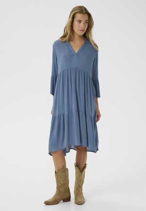 Abito blu, fino al ginocchio, con scollatura a V, maniche a campana e design a strati; abbinato a stivali da cowboy color nocciola con cuciture decorative.