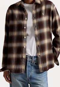 Brun och beige rutigt flanellskjorta med knappar, långa ärmar och rundad nederkant. Bärs över en vit t-shirt och blå jeans.