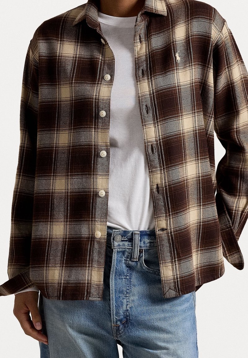 Brun och beige rutigt flanellskjorta med knappar, långa ärmar och rundad nederkant. Bärs över en vit t-shirt och blå jeans.