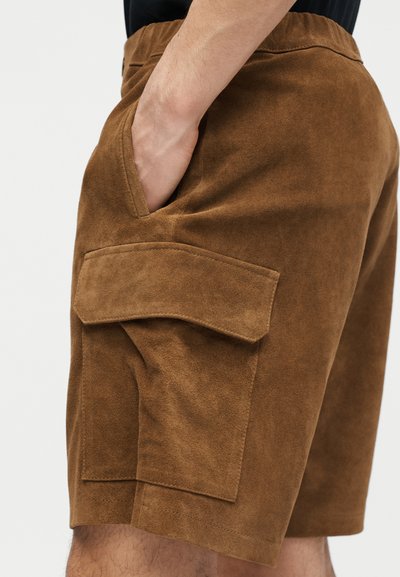 Shorts cargo en suède marron avec grande poche latérale, portés par une personne ayant la main dans la poche avant, sur un fond uni.