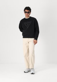 Sudadera negra con un logotipo de triángulo, combinada con pantalones beige claros y zapatillas grises. El tejido parece suave; el aspecto es casual.