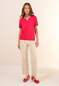 Chemise rouge à manches courtes avec un col en V, associée à un pantalon large beige et des baskets rouges avec des lacets blancs. Tenue simple et décontractée.