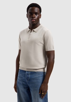 Young man wearing a beige short-sleeve polo shirt with a zipper and blue jeans, standing against a plain light background.