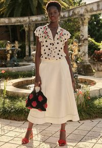 Blusa crema con pois neri e ricami floreali, abbinata a una gonna crema. Accentuata con una borsa nera decorata con rose rosse e sandali con tacco rossi.