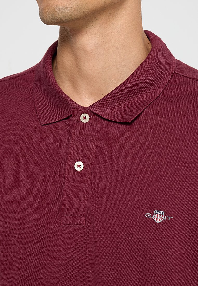 Polo bordeaux avec un col côtelé, une patte de boutonnage à deux boutons et le logo GANT brodé sur la poitrine. Le tissu semble texturé et respirant.