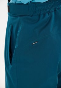 Türkisblaue Hose mit seitlichem Reißverschluss und kleinem Whistler-Label an der Oberseite des Oberschenkels, getragen von einer Person in einem passenden blauen Hemd.