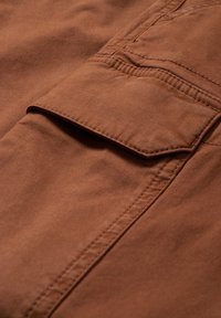 Gros plan sur une poche cargo en tissu marron avec rabat et coutures visibles sur un vêtement.