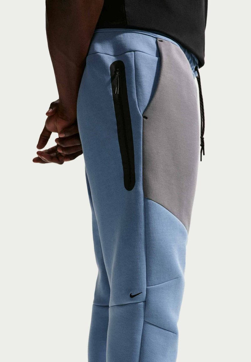 Pantaloni jogger azzurri realizzati in tessuto morbido con pannelli grigi. Presentano tasche con cerniera e dettagli neri, incluso un piccolo logo.