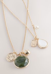 Gold-tone layered necklace featuring multi-shaped pendants: a green stone, a pearlescent drop, a textured gold charm, and a flat circular piece.