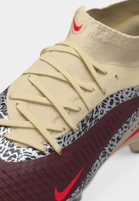 Μπεζ και μαρόν ποδοσφαιρικό παπούτσι Nike με κίτρινες κορδόνια και ασπρόμαυρο πουά μοτίβο κοντά στη σόλα, με κόκκινους λογότυπους σβούρας.