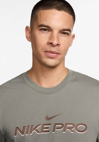 Camiseta Nike Pro gris de manga corta con un atrevido diseño de logo y franjas marrones en el pecho. Material de algodón suave, cuello redondo.