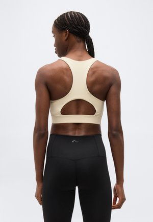 Kobieta z zaplecionymi włosami, ubrana w beżowy sportowy top na ramiączkach w stylu racerback i czarne legginsy z wysokim stanem, stojąca tyłem do kamery.