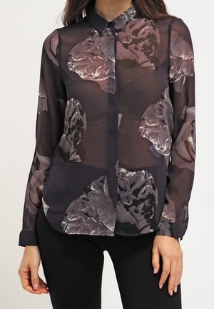 Kvinde iført en sort gennemsigtig skjorte med knapper og store grå blomsterprint samt sorte bukser, stående med afslappede arme.