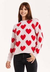Light pink sweater with raised red heart patterns. Crew neckline, long sleeves, textured fabric. Paired with black jeans.