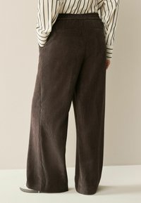 Braune Corduroy-Hose im Weitschnitt mit strukturiertem Stoff, elastischem Bund und Seitentaschen. Das Kleidungsstück wird mit einem gestreiften Hemd kombiniert.