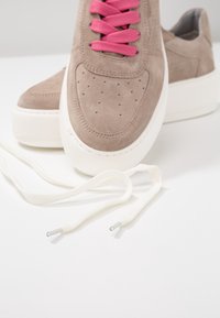 Baskets en daim taupe avec des lacets rose vif et une semelle blanche épaisse, avec une paire supplémentaire de lacets blancs posée à proximité sur une surface blanche.