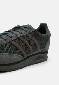 Ciemnoszary but sportowy Adidas z dzianinowym materiałem, zamszowymi nakładkami, trzema brązowymi paskami po boku i teksturowaną gumową podeszwą.