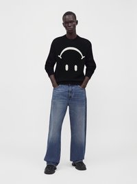 Homme portant un pull noir avec un motif de visage souriant blanc, jean bleu ample et chaussures noires, debout les mains dans les poches sur fond blanc.