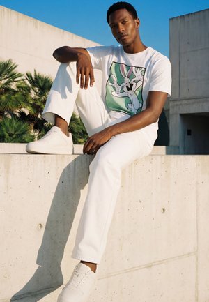 Mladić u bijelim tenisicama, hlačama i majici s likom Bugs Bunnyja sjedi na betonskom zidu na otvorenom uz palme i plavo nebo.